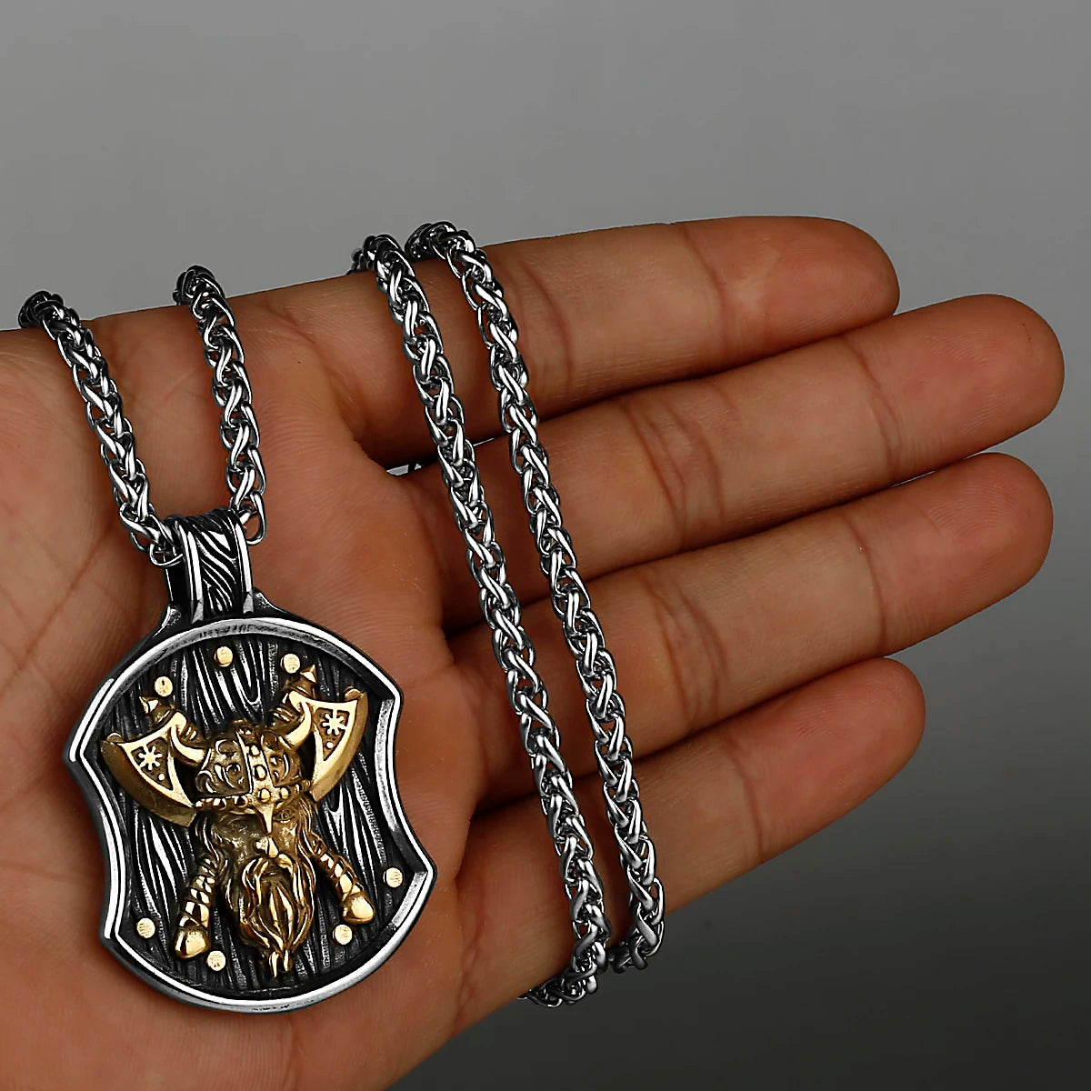 A hand holds a stainless steel Viking warrior shield necklace with a chain and pendant.