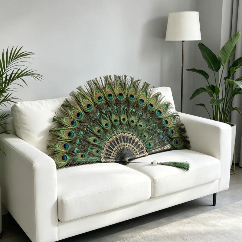 Un éventail à plumes de paon naturel tenu à la main, exposé sur un canapé blanc dans un salon moderne.