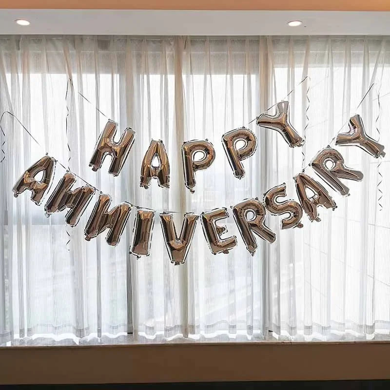 A silver foil balloon banner spelling out Happy Anniversary strung across a window with sheer white curtains.