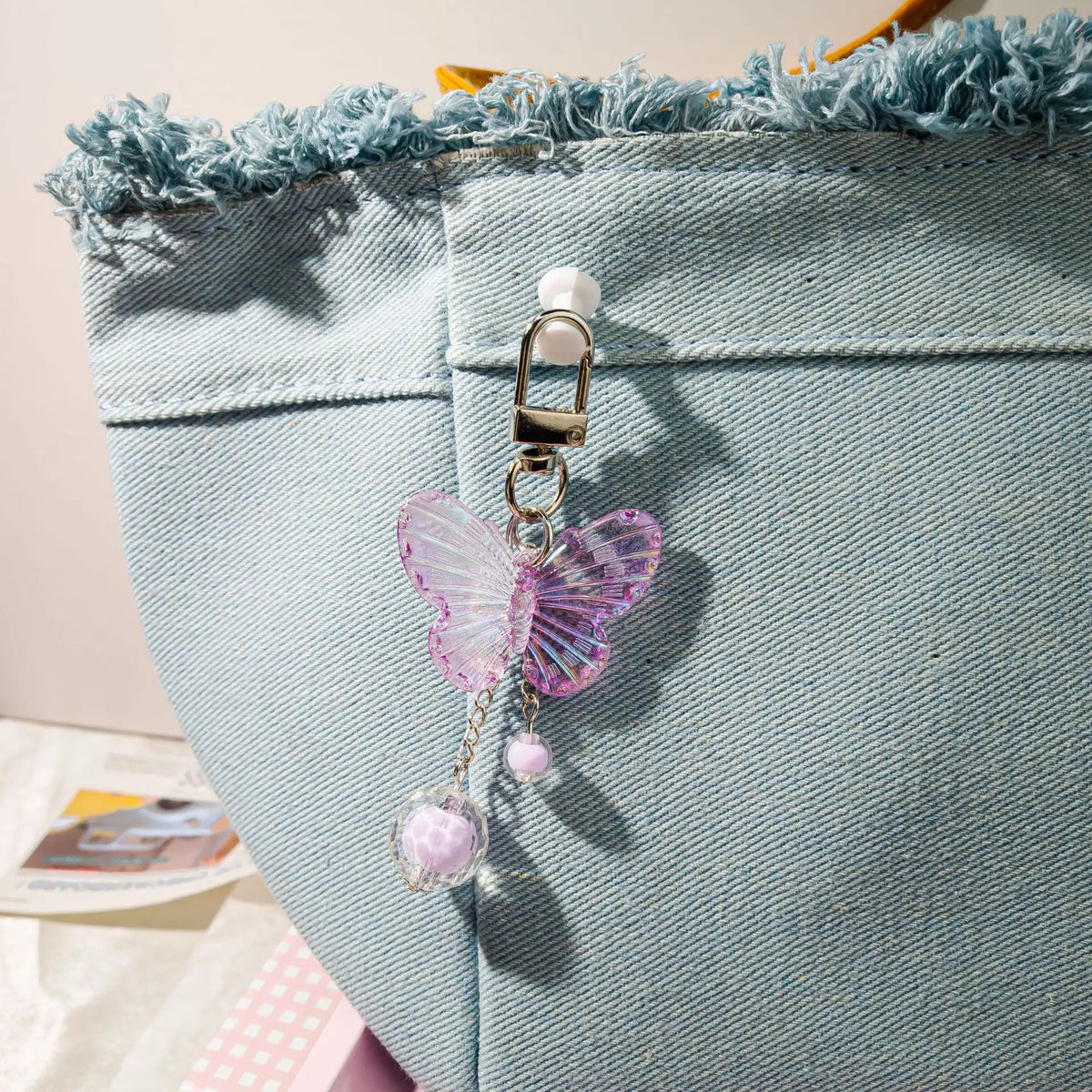 A pink butterfly pendant keychain set with pearl beads hanging from a silver clasp against a light blue fabric background.