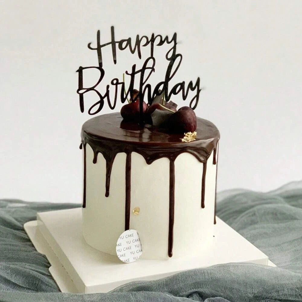 A white cake with chocolate drip and gold lettering spelling out Happy Birthday, set on a light platform.