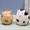 A cute, light brown cow piggy bank with pink accents and a black and white pattern sitting on a light surface.