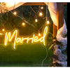 A glowing neon sign spelling Just Married is displayed outdoors with greenery and warm string lights.
