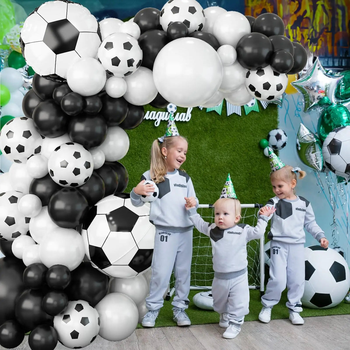 A family of three children playing with a large black and white soccer ball in front of a green backdrop decorated with ballo