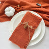 A stack of ten handcrafted fringed cotton napkins in a rust orange color resting on white plates and an orange textile.