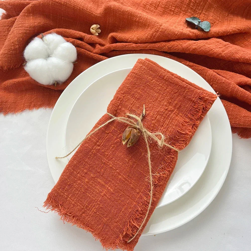 A stack of ten handcrafted fringed cotton napkins in a rust orange color resting on white plates and an orange textile.