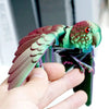 A hand holds an articulated 3D printed parrot figurine with movable wings, featuring green and reddish-pink plumage.