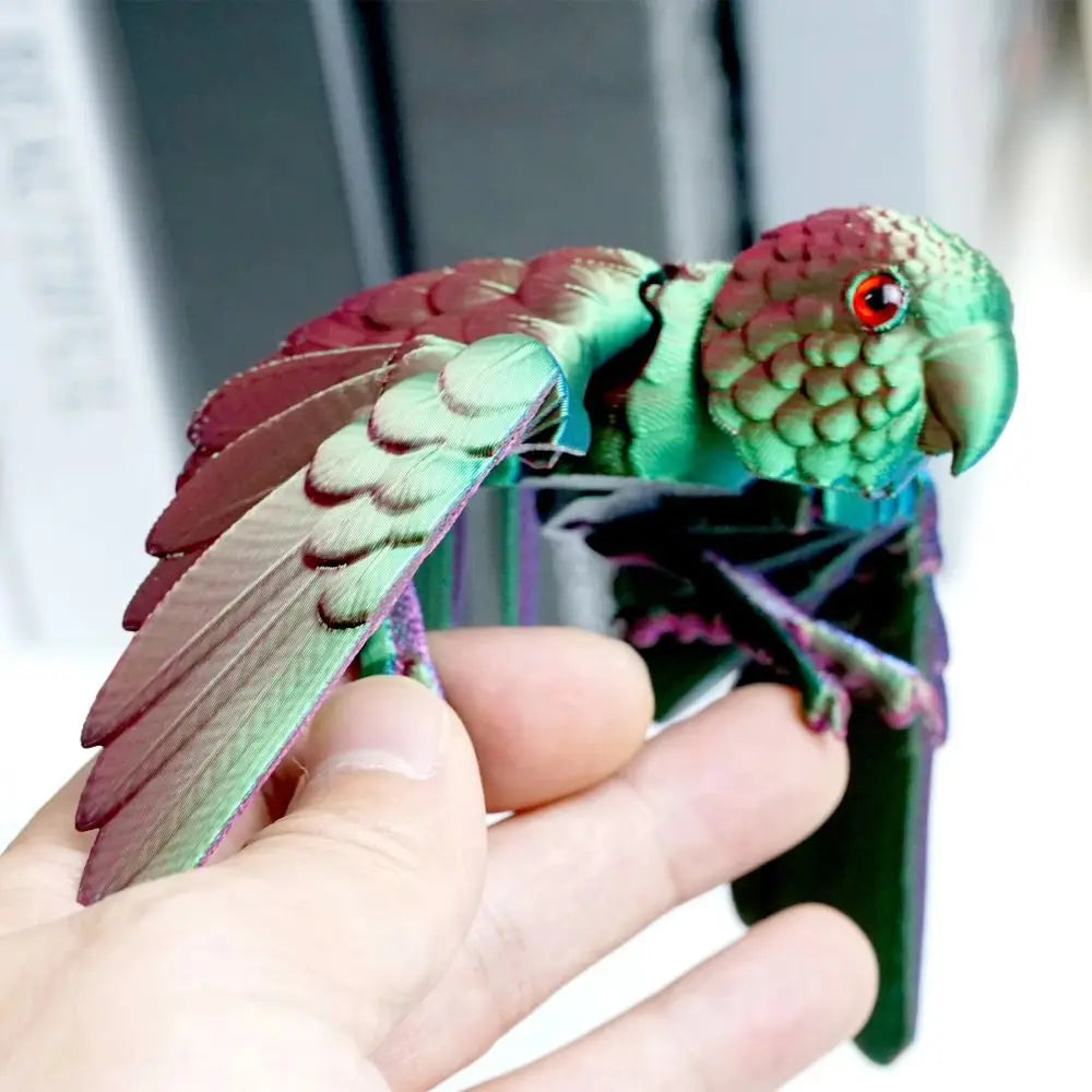 A hand holds an articulated 3D printed parrot figurine with movable wings, featuring green and reddish-pink plumage.