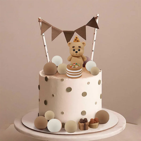 A round cake decorated with polka dots and topped by a teddy bear figure and triangular pennant banners.