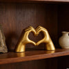 A golden sculpture of two hands forming a heart, displayed on a wooden shelf.