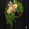A rustic green wreath featuring cream hydrangeas and a burlap bow, mounted on a dark door.