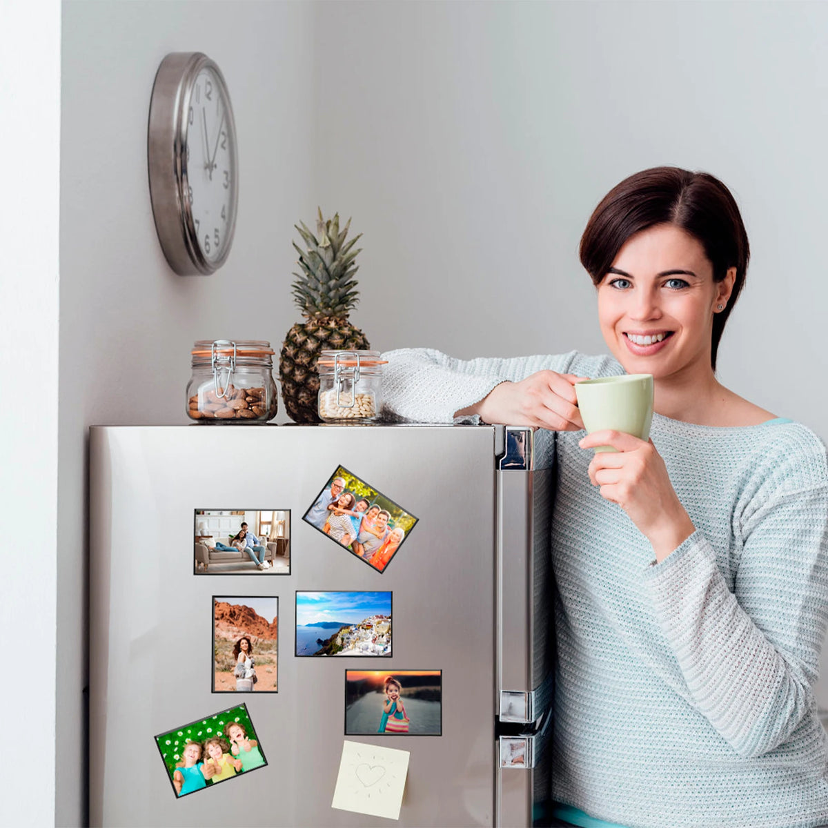 A woman holds a mug in front of a refrigerator decorated with magnetic picture frames.
