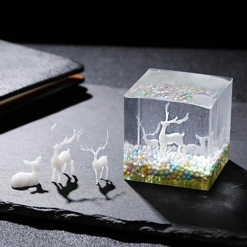 A clear resin block filled with colorful glitter and white deer figurines rests on a dark surface.