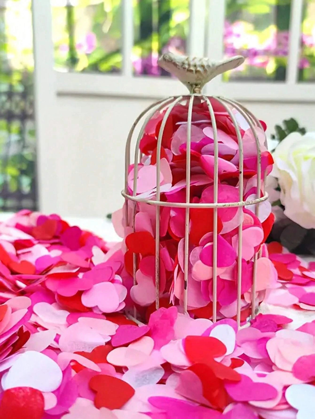 A silver cage filled with pink and red heart sequins confetti rests on a surface covered in loose petals.