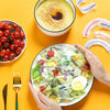 Hands holding a salad in a plastic food cover next to a bowl of soup and fresh tomatoes on a yellow background.