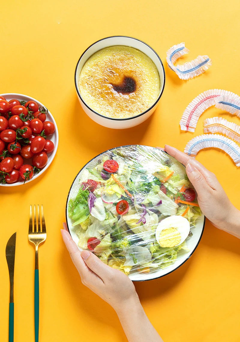 Hands holding a salad in a plastic food cover next to a bowl of soup and fresh tomatoes on a yellow background.