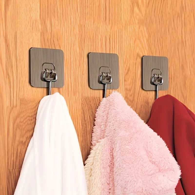 Three silver adhesive wall hooks mounted on a wooden surface, with various colored fabric items hanging below.