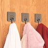 Three silver adhesive wall hooks mounted on a wooden surface, with various colored fabric items hanging below.