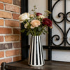 A floral arrangement of roses in a black and white striped ceramic vase sits on a dark wooden surface next to a brick wall an