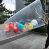 A giant clear plastic bag filled with colorful balloons is being held by a person outdoors.