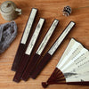 A set of dark wood folding fans with white paper and bamboo elements resting on a wooden surface next to a pinecone.