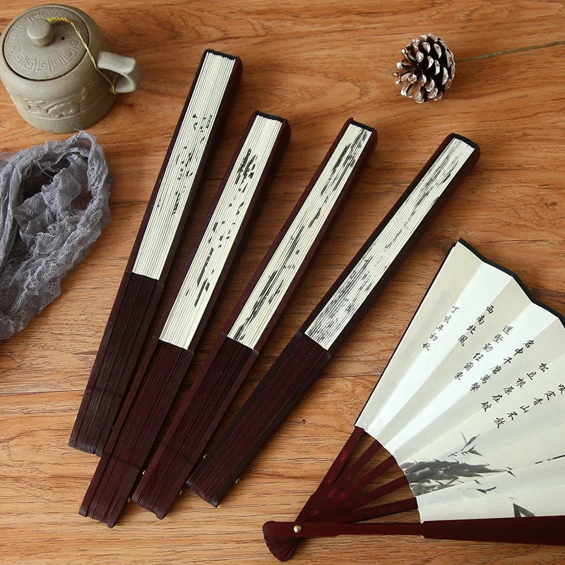 A set of dark wood folding fans with white paper and bamboo elements resting on a wooden surface next to a pinecone.