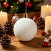 A white velvet flocked Christmas ornament is displayed on a wooden surface with pine cones and festive lights in the backgrou
