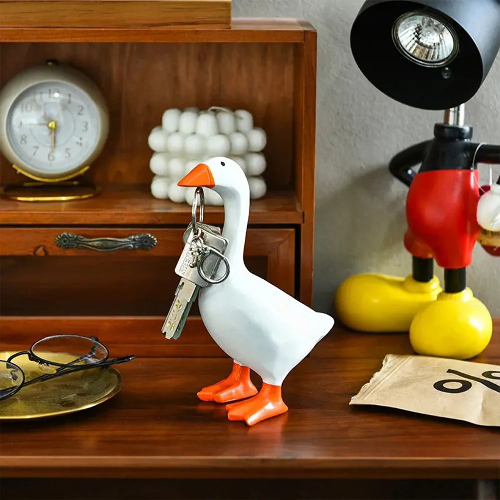 A white ceramic goose figurine with orange feet and a key holder is displayed on a wooden table next to a lamp.