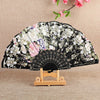 A black silk flower folding fan decorated with white and pink floral patterns, displayed on a wooden stand.
