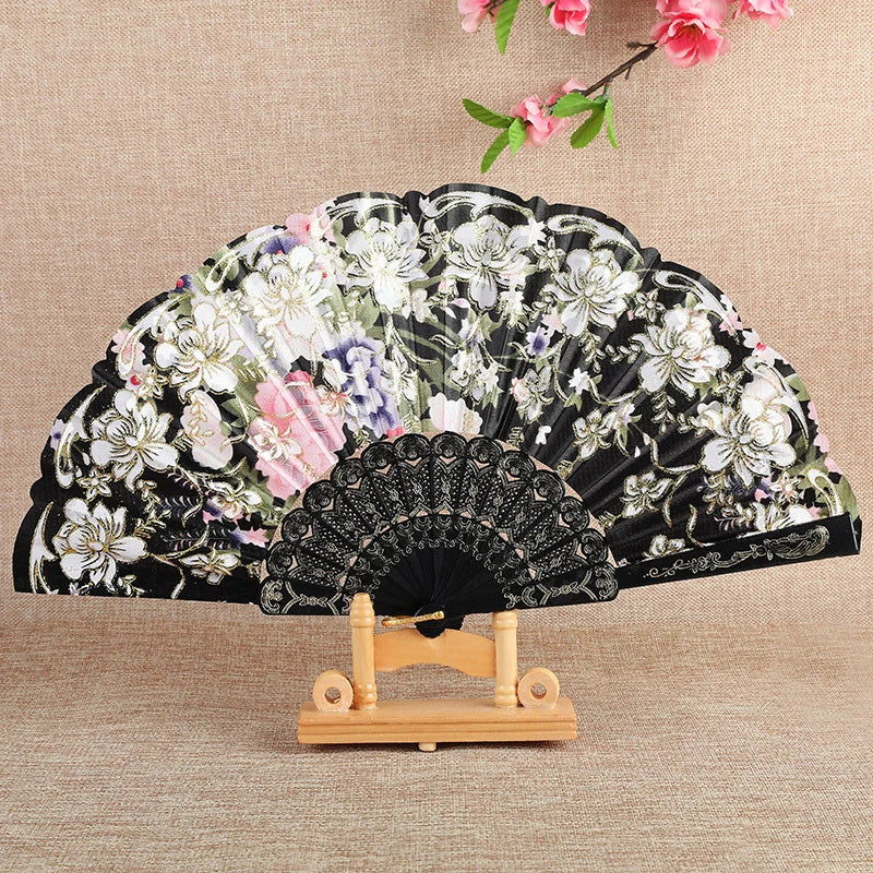 A black silk flower folding fan decorated with white and pink floral patterns, displayed on a wooden stand.