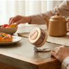 A hand is operating a magnetic bladeless handheld fan on a wooden surface near a teapot and food.