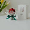 A red tulip bouquet wrapped in sheer fabric and tied with a green ribbon next to its white candle box.