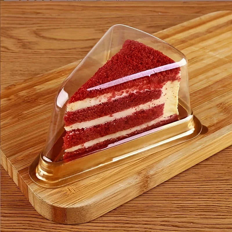 A slice of red velvet cake layered with cream is displayed in a clear disposable plastic container on a wooden board.