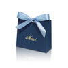 A dark blue paperboard gift bag tied with a light blue ribbon, featuring the word Merci written in gold script.