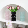 A display of handcrafted glass flower vases in shades of pink and green on a white shelf.