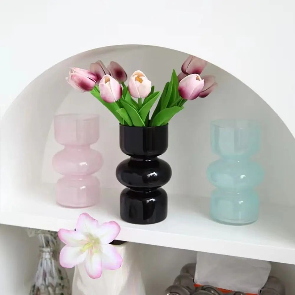 A display of handcrafted glass flower vases in shades of pink and green on a white shelf.