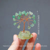 A hand holds a natural crystal tree made of green stones branching out from a larger crystal base.