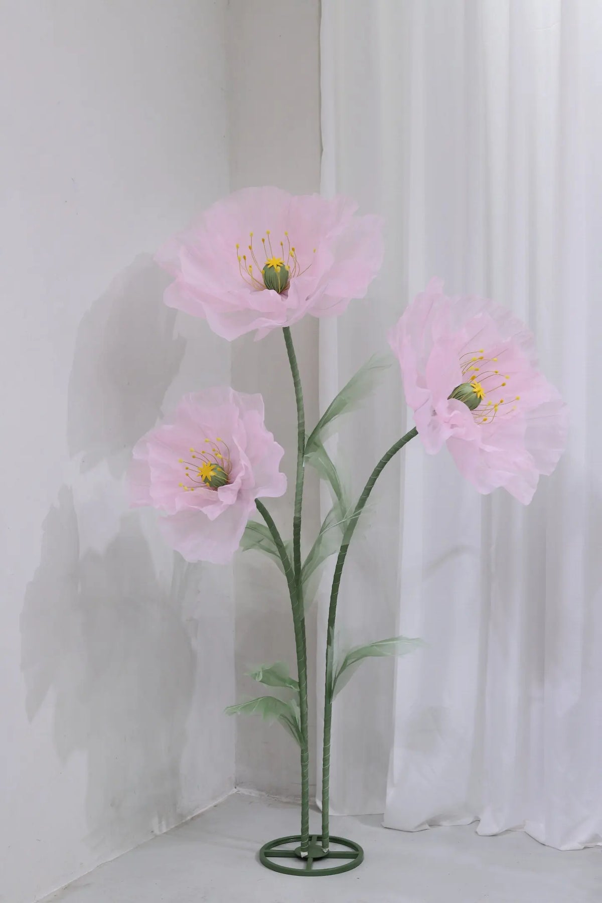 A large artificial silk pink poppy flower with yellow stamens displayed on a green stand against a white background.