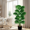 A large artificial palm tree in a black pot placed indoors near a window with sheer curtains.