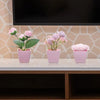A set of three pink artificial rose potted plants displayed on a white table against a light tiled background.