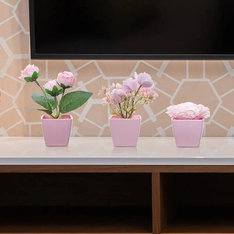 A set of three pink artificial rose potted plants displayed on a white table against a light tiled background.