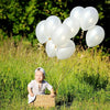 A young child sits in a grassy field holding strings of white latex balloons.