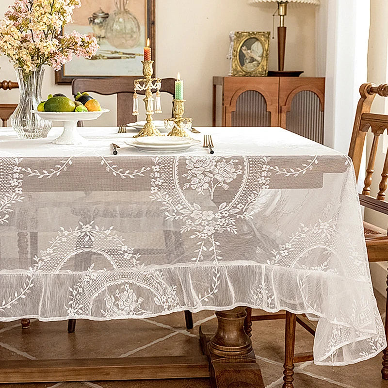 Elegant white floral lace tablecloth spread on a wooden table with candles and decorative items.