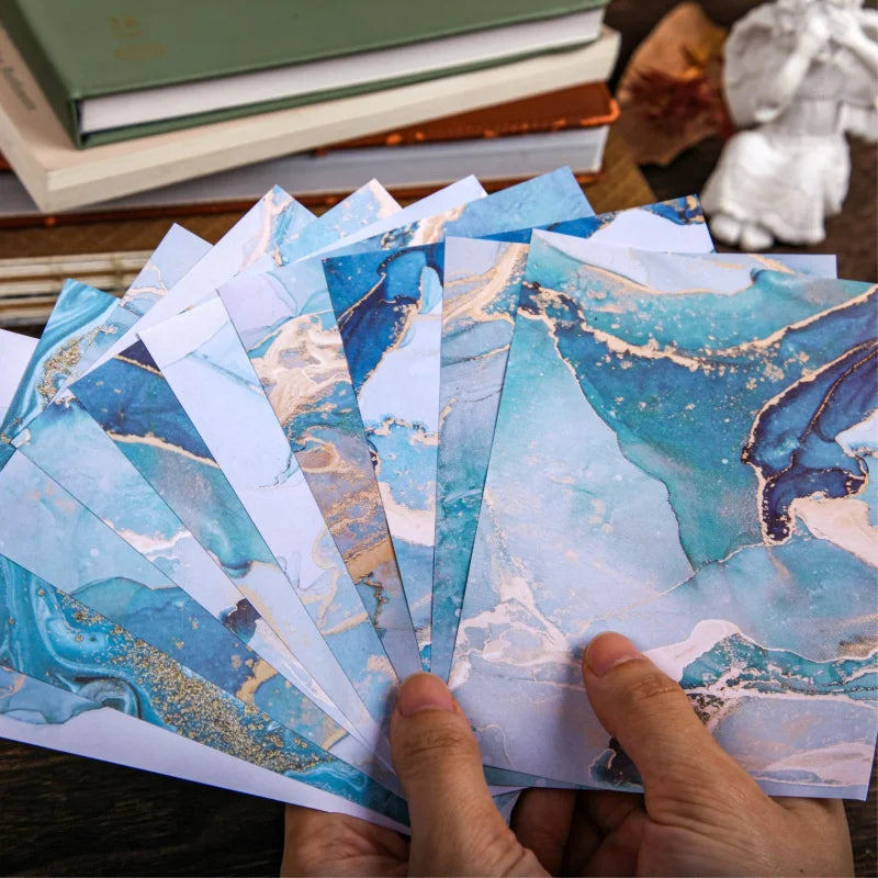 A hand holds a stack of gradient wash retro decorative paper sheets featuring blue and gold marbled textures.