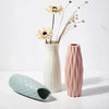 A tall, pale pink vase holds dried white flowers and thin stems on a white surface.
