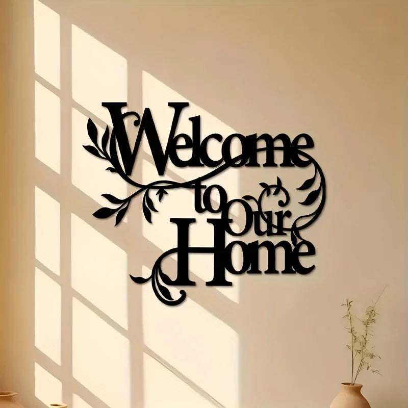 A black metal welcome sign with the text Welcome to Our Home mounted on a light wall, casting shadows.