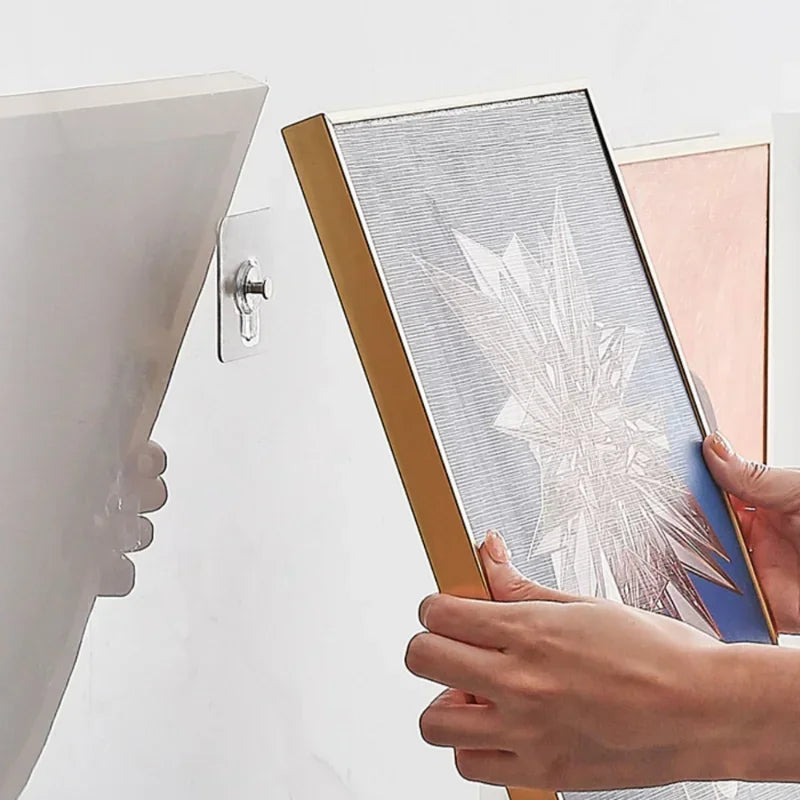 Hands holding a framed picture with a gold frame and silver-toned artwork against a white wall.