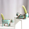 Two ceramic feline-shaped succulent planters with fuzzy green plants sitting on a white surface.