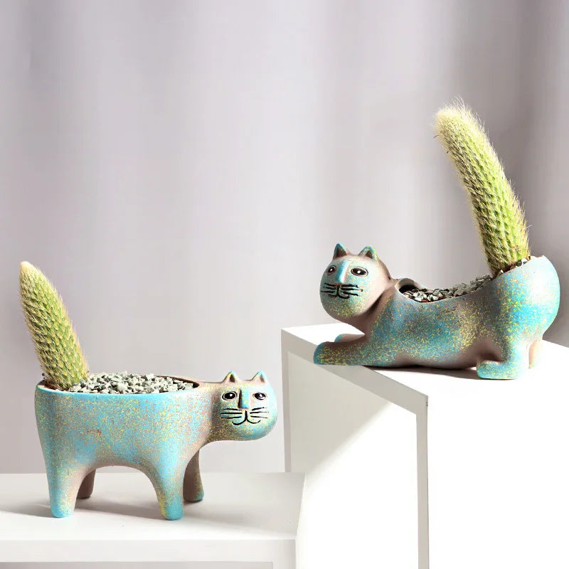 Two ceramic feline-shaped succulent planters with fuzzy green plants sitting on a white surface.