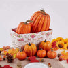 A set of seven orange artificial pumpkins arranged in a woven basket on a table with autumn leaves and flowers.